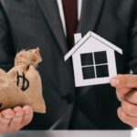 Homme qui tient une petite maison en papier dans une main et deux sacs d'argent dans l'autre